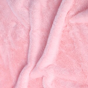 May include: Close-up of a soft, pink, faux fur fabric. The plush material is folded and draped, creating shadows and highlights that emphasize its texture. The color is a light, pastel pink, suggesting a cozy and comfortable item.