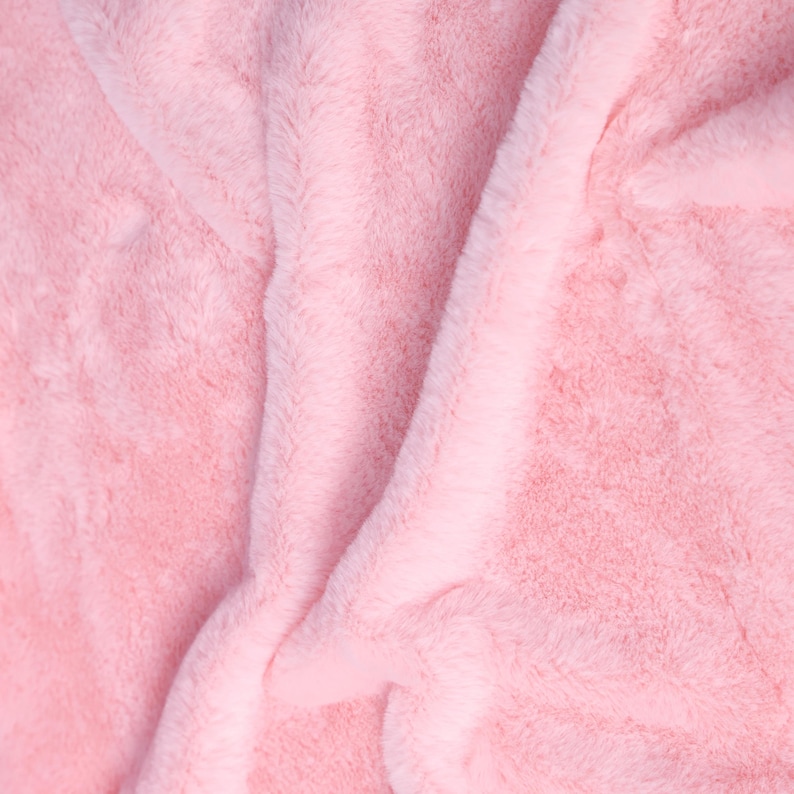 May include: Close-up of a soft, pink, faux fur fabric. The plush material is folded and draped, creating shadows and highlights that emphasize its texture. The color is a light, pastel pink, suggesting a cozy and comfortable item.