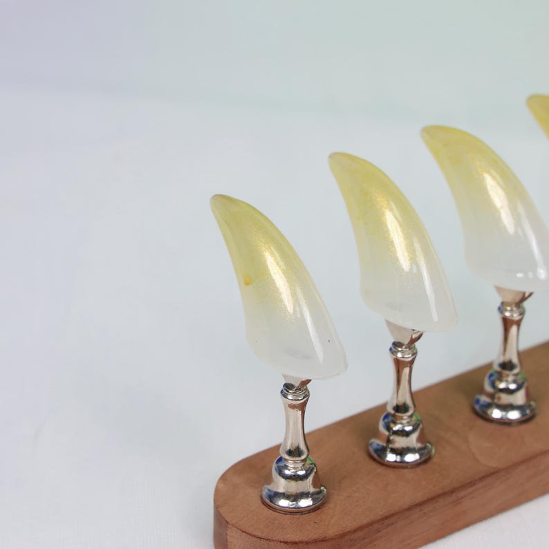 May include: Four decorative, claw-shaped objects with a gradient of yellow to white. Each claw is mounted on a silver-colored pedestal and set on a wooden base. The background is a plain white.