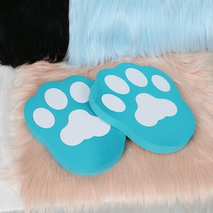 May include: Two turquoise paw-shaped cushions with white paw print details. The cushions are placed on a pink, fluffy surface, with black and light blue faux fur in the background. These cushions are designed for comfort and style.