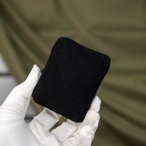 May include: A small, rectangular, solid black fabric pouch with a fine-knit texture. The item features rounded corners and a consistent, matte black finish across its entire visible surface, presenting a simple, uniform, and compact design.