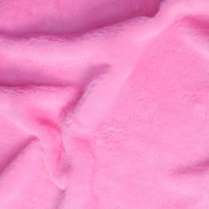 May include: Close-up of a vibrant pink faux fur fabric. The plush material has a soft, textured appearance with visible fibers and subtle variations in color, creating a luxurious look. The fabric is folded, showing depth and dimension.
