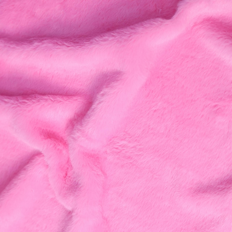 May include: Close-up of a vibrant pink faux fur fabric. The plush material has a soft, textured appearance with visible fibers and subtle variations in color, creating a luxurious look. The fabric is folded, showing depth and dimension.