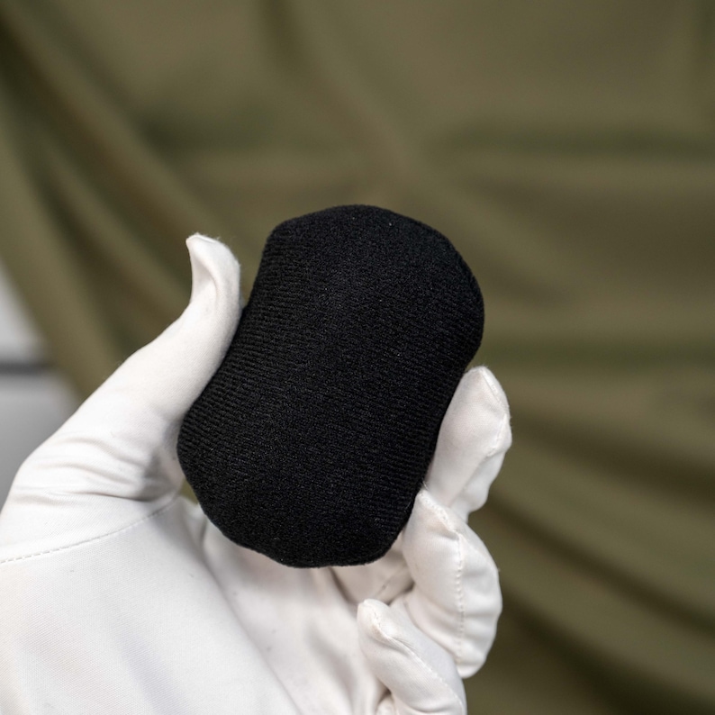May include: A small, solid black, oval-shaped object with a textured, fabric-like surface. The item features a rounded, pill-like form with a consistent matte finish across its entire exterior, showing a dense, woven material construction.
