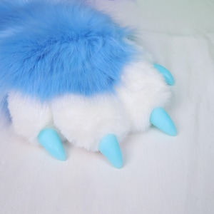 Op de afbeelding: Een harige poot in blauw en wit, met lichtblauwe klauwen. Het pluche materiaal heeft een zachte textuur. De poot maakt waarschijnlijk deel uit van een kostuum of een pluche speelgoed.