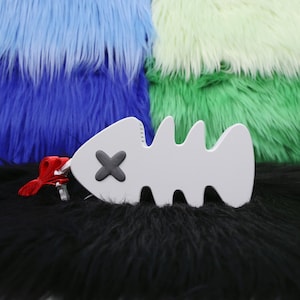 May include: A white, fish-shaped accessory with a black X on one side and a red tassel. The accessory is set against a backdrop of blue, green, and black faux fur.