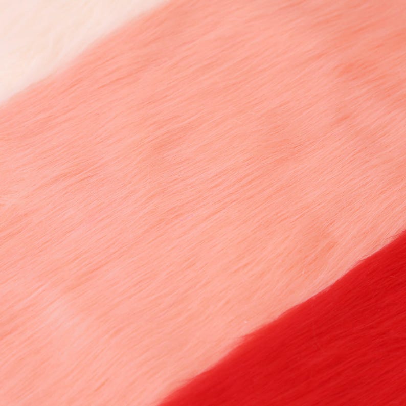 May include: Close-up of a colorful faux fur fabric. The image features a diagonal color block design with sections of white, light pink, medium pink, and red. The soft texture of the fur is visible.