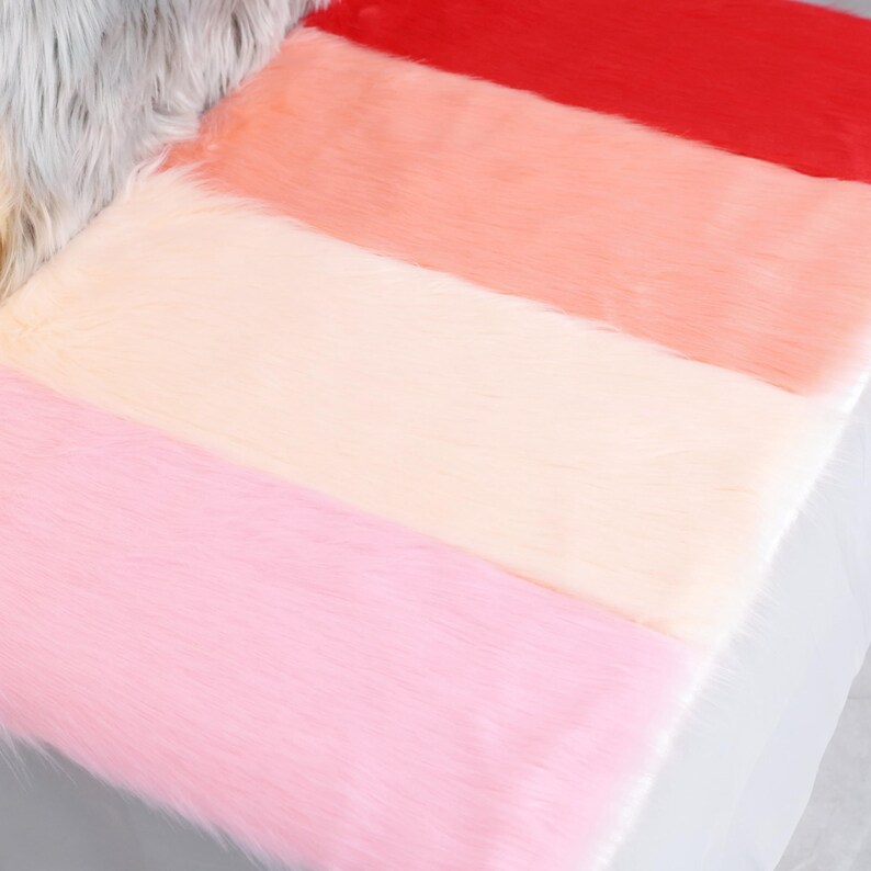 May include: A close-up of a faux fur fabric in a gradient of colors. The colors include red, coral, peach, cream, and pink. The fur is soft and fluffy, with long strands. The fabric is likely used for crafts or decor.