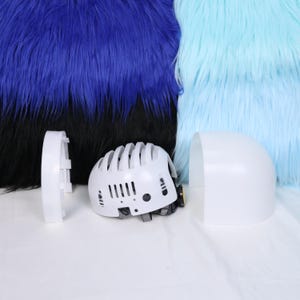 Może przedstawiać: Biały, zdemontowany kask z czarnym paskiem pod brodą i małym żółtym światłem. Kask znajduje się obok białego, zakrzywionego elementu i białego, prostokątnego elementu. Tło przedstawia niebieskie i jasnoniebieskie sztuczne futro.