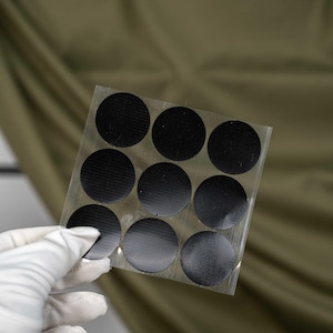 May include: A square transparent sheet containing nine circular black adhesive patches arranged in a three-by-three grid. Each patch features a fine, textured, cross-hatched surface pattern, designed for repair or reinforcement applications.