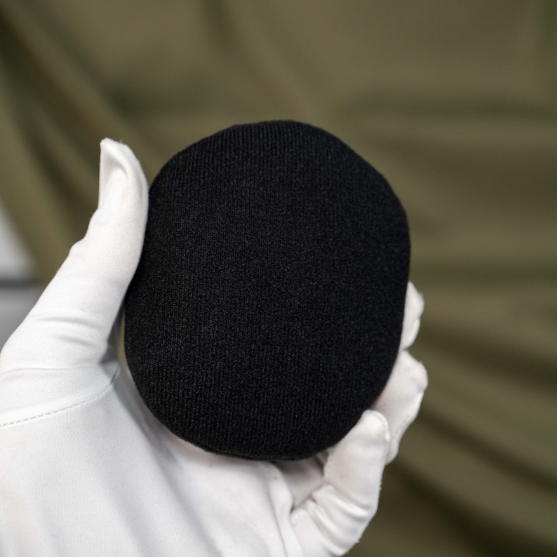 May include: A solid black, circular, fabric-covered object with a smooth, fine-knit texture. The item is rounded and compact, featuring a consistent dark color throughout its surface, presenting a simple, uniform, and clean appearance.