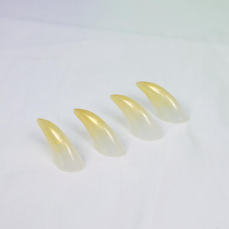 May include: Four translucent, claw-shaped objects with a gradient of yellow to clear. The objects are arranged diagonally on a white surface, suggesting a set of costume or decorative pieces.