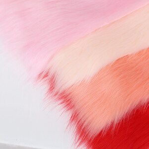 May include: Close-up of faux fur fabric in a gradient of pink and red hues. The colors transition from light pink to a deeper red, creating a soft, textured appearance. The fur fibers are long and fluffy, suggesting a plush material.