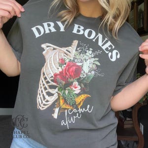 May include: Dark gray t-shirt with the words "DRY BONES" above a graphic of a rib cage with a floral arrangement and the words "come alive" in script. The floral arrangement includes red roses and a butterfly.