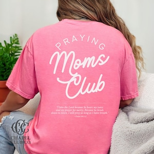 May include: A pink t-shirt with the words "PRAYING Moms Club" in white script on the back. Below the text is a Bible verse. The Chapel Club Co. logo is visible on the bottom left.