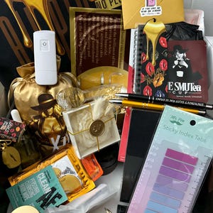 May include: An open box filled with various items, including a white device, a gold-colored statue, a notebook with red lips, pens, sticky index tabs, and skincare products. The box has a black background with gold dripping liquid.