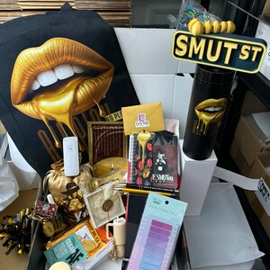 May include: A black tote bag and a black tumbler with gold lips design, a sign that says "SMUT ST", and a box filled with various items, including a notebook, sticky index tabs, and a small gold bag. The items are arranged on a table.