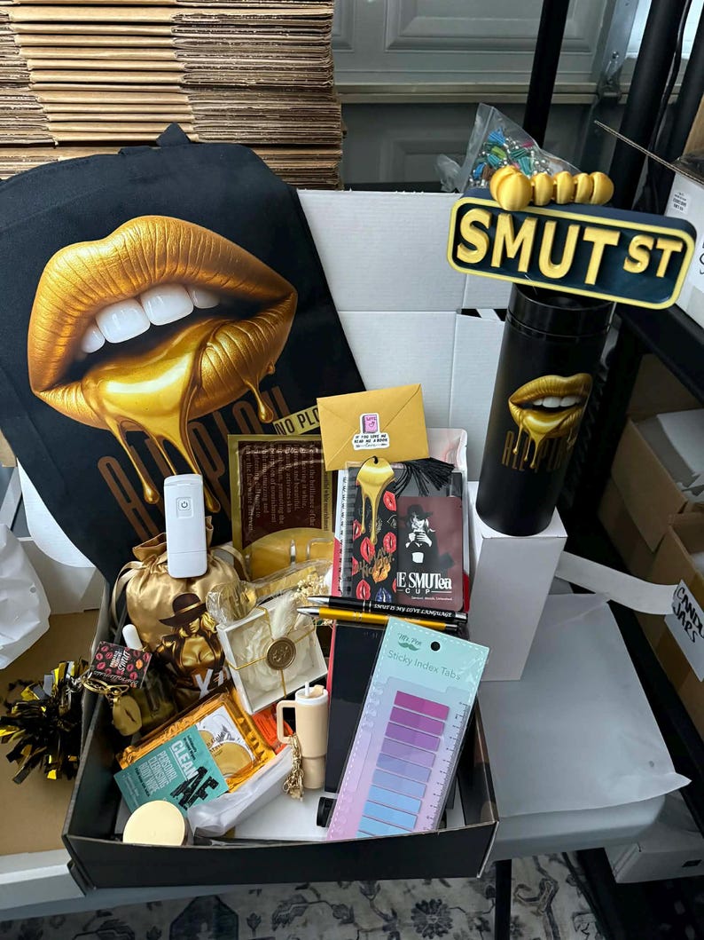 May include: A black tote bag and a black tumbler with gold lips design, a sign that says "SMUT ST", and a box filled with various items, including a notebook, sticky index tabs, and a small gold bag. The items are arranged on a table.