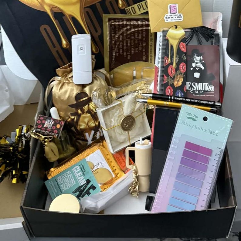 May include: A gift box filled with various items, including a white electronic device, a gold-colored bag, a small mug, a pen, and sticky index tabs. The box also contains a small envelope and a card with the text "ESMUTEA CUP".
