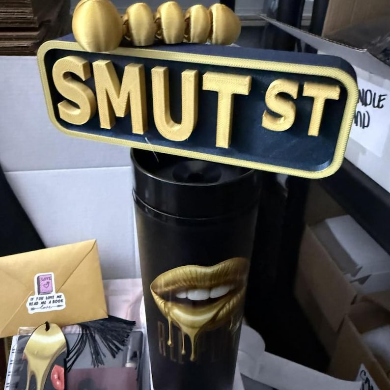 May include: A black tumbler with a gold lip design and the word "RATED". Above it, a sign that reads "SMUT ST" in gold letters on a navy blue background. Several gold rings are on top of the sign. A gold bookmark and a small envelope are also visible.
