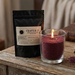 Puede incluir: Una vela encendida en un vaso transparente con cera granular de color rojo intenso. A la izquierda hay una bolsa negra con una etiqueta blanca que dice "CHAPTER 13". La vela y la bolsa están sobre una superficie de madera. La vela forma parte de la "SMUT BOX COLLECTION".
