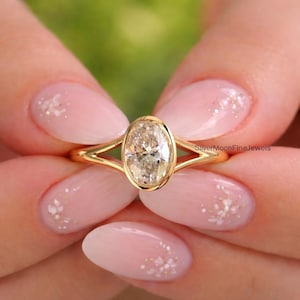 May include: A gold ring featuring an oval-shaped centre stone set in a bezel setting with a split shank band. The ring includes the text SilverMoonFineJewels printed on the side of the band. The design showcases a polished yellow gold finish.