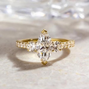 Könnte beinhalten: Ein goldener Verlobungsring mit einem großen Diamanten im Marquiseschliff als Mittelstein, flankiert von zwei kleineren runden Diamanten. Das Band ist ebenfalls mit kleineren Diamanten besetzt, was einen funkelnden Effekt erzeugt. Der Ring liegt auf einer hellen Oberfläche.