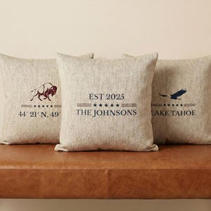 Könnte beinhalten: Drei quadratische Zierkissen mit neutraler Leinenstruktur. Ein Kissen zeigt eine Bison-Grafik und Koordinaten, ein anderes lautet "EST 2025 THE JOHNSONS", und das dritte zeigt einen fliegenden Adler und "LAKE TAHOE".