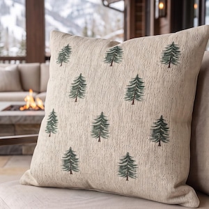 May include: A square throw pillow featuring a textured beige fabric base with a repeating pattern of small, dark green pine trees. The pillow has a visible woven grain and is positioned on a neutral-colored outdoor chair cushion.