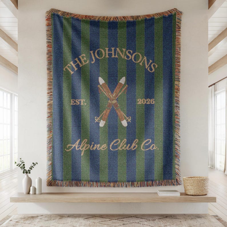 May include: A large, rectangular throw blanket with vertical blue and green stripes. The blanket features the text "THE JOHNSONS" above crossed skis, with "EST. 2026" and "Alpine Club Co." in a cursive font. The edges have a fringed trim.