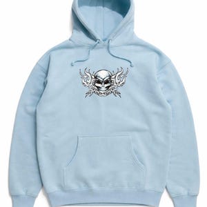 Puede incluir: Sudadera con capucha azul claro con un diseño de calavera y llamas. La calavera es en blanco y negro con detalles rojos en los ojos. La sudadera tiene un bolsillo delantero y una capucha con cordón.
