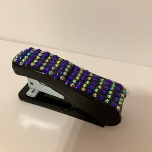 May include: Black stapler bedazzled with iridescent purple and blue rhinestones. The stapler has a silver metal base and a black handle. The rhinestones are arranged in a grid pattern across the top of the stapler.