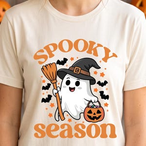 May include: A cream-coloured t-shirt featuring a graphic of a white ghost wearing a black witch hat, holding a broomstick and a jack-o'-lantern candy bucket. The text "SPOOKY season" is printed in orange above and below the ghost illustration.