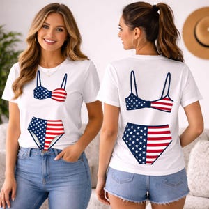 May include: A white short-sleeve t-shirt featuring a graphic print of a bikini with a red, white, and blue American flag pattern. The design includes a bra top and high-waisted bottoms, displayed on the front and back of the garment.