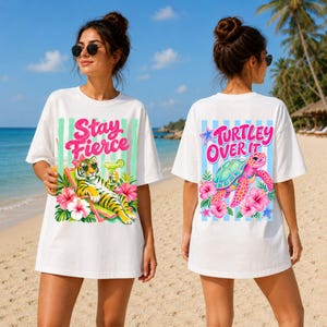 Op de afbeelding: Witte T-shirts met grafische ontwerpen. Eén shirt heeft een tijger met een zonnebril en de tekst "Stay Fierce". Het andere shirt heeft een schildpadontwerp met de tekst "Turtley Over It". Beide shirts hebben een strandthema.