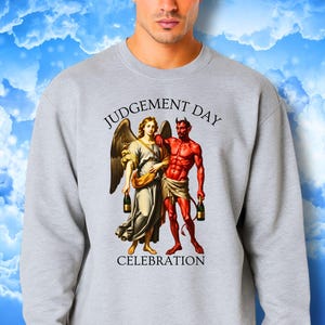May include: A gray sweatshirt featuring a graphic of an angel and a devil holding bottles, with the words "JUDGEMENT DAY CELEBRATION" in black. The angel is in a cream-colored dress, and the devil is red.