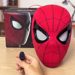 May include: A red Spider-Man mask featuring black web patterns and white mesh eye lenses, accompanied by a small black remote control with five buttons. The mask is positioned in front of its black product packaging box with matching graphics.