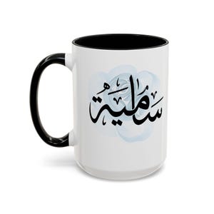 May include: White ceramic mug with a black handle and interior. The mug features a black Arabic calligraphy design over a light blue floral background. The mug is a standard size for coffee or tea.