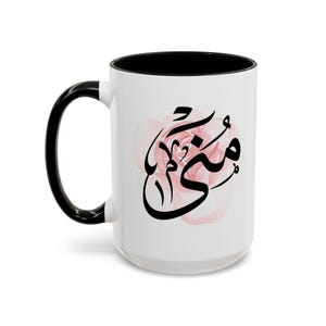 May include: White ceramic mug with a black handle and interior. The mug features a black Arabic calligraphy design over a soft pink floral background. The mug is a standard size for coffee or tea.