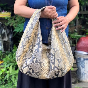 Peut inclure: Un grand sac à main souple avec un motif peau de serpent dans les tons beige, gris et noir. Le sac est tenu par les anses et semble être fait d'une matière souple et pliable. Le design du sac suggère un style décontracté et quotidien.