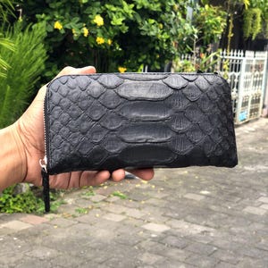 May include: Black wallet with a textured, scaly pattern, held in a hand. The wallet has a zipper closure and a wrist strap. The background shows greenery and a paved surface.