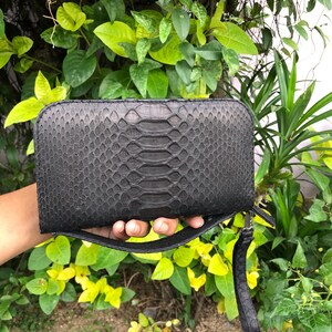 K&ouml;nnte beinhalten: Eine schwarze, rechteckige Clutch mit Schlangenmuster. Die Clutch hat eine Handschlaufe und einen Rei&szlig;verschluss. Der Hintergrund zeigt gr&uuml;nes Laub und gelbe Bl&auml;tter.
