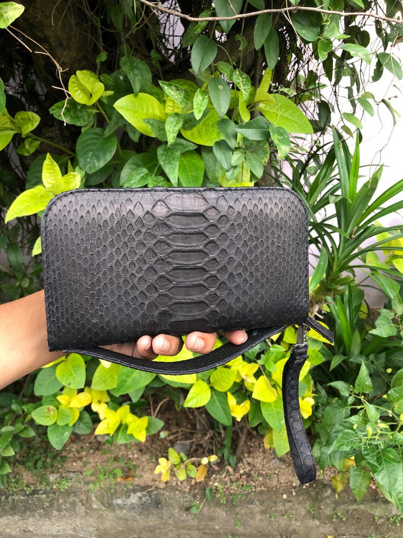 K&ouml;nnte beinhalten: Eine schwarze, rechteckige Clutch mit Schlangenmuster. Die Clutch hat eine Handschlaufe und einen Rei&szlig;verschluss. Der Hintergrund zeigt gr&uuml;nes Laub und gelbe Bl&auml;tter.