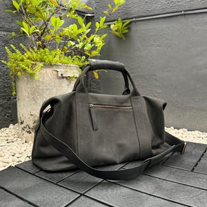 Peut inclure: Un sac de voyage en cuir gris foncé avec une poignée supérieure et une bandoulière amovible. Le sac a une poche avant zippée et une surface texturée. Le sac est posé sur une surface noire.