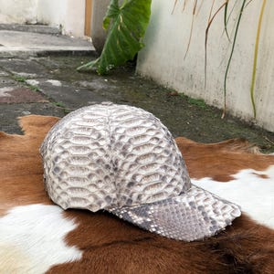 May include: A baseball cap featuring a textured, scale-like pattern in shades of off-white, beige, and grey. The cap has a rounded crown and a curved brim, with the material displaying a distinct, repeating organic pattern across the entire surface.