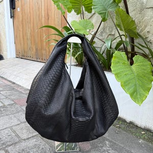 May include: A large, black leather shoulder bag with a snakeskin texture. The bag has a single strap and a large, open interior. The bag is displayed on a silver stand, with green plants in the background.