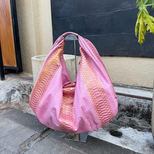 Pode incluir: Uma bolsa rosa com um padrão de pele de cobra em dourado e rosa. A bolsa é exibida em um suporte prateado. A bolsa tem um design grande e aberto, e o material parece macio e flexível. O fundo inclui uma superfície de concreto e uma parede escura.