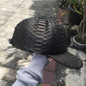 May include: A black baseball cap with a textured, scaly pattern. The cap has a curved brim and a strap in the back for adjustment. The cap is made of a dark material, possibly leather or a similar material.