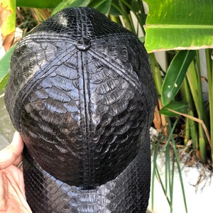 May include: A black baseball cap featuring a textured, scale-like pattern across the entire surface. The cap is constructed with six panels that meet at a central button on the crown, and it includes a curved brim, all finished in a glossy black.