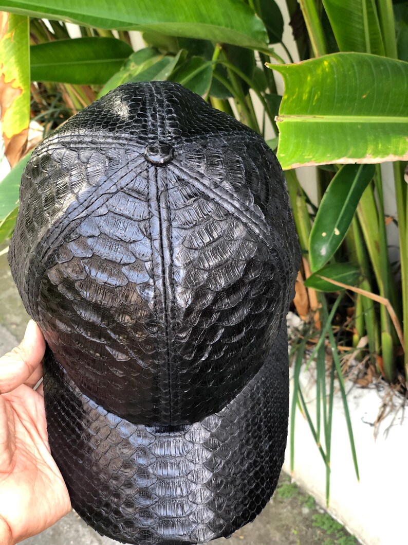 May include: A black baseball cap featuring a textured, scale-like pattern across the entire surface. The cap is constructed with six panels that meet at a central button on the crown, and it includes a curved brim, all finished in a glossy black.